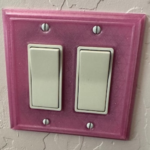 Puede incluir: Cubierta de interruptor de luz rectangular rosa con dos interruptores basculantes. La cubierta está fijada a la pared con cuatro tornillos. Los interruptores son de color blanquecino.
