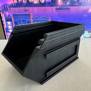 May include: A black, angular container with a stepped design on one side and a rectangular panel on the other. The container is made of a matte black material and has a geometric shape. The background features a colorful cityscape.