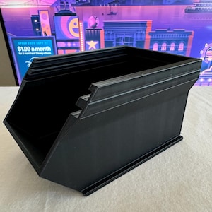 May include: A black, angular storage container with a stepped design on the top edge. The container has a rectangular base and a sloped front. The background includes a promotional graphic with the text "OFFER ENDS SEPT 27 $1.99 a month for 3 months of Disney+ Basic."