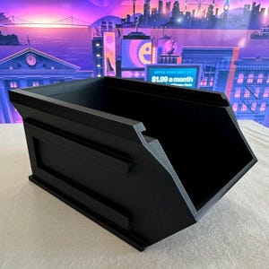 May include: A black, angular storage container with a rectangular front panel and two horizontal bars. The container has a sloped design and a wide opening. The background features a cityscape with a purple and blue color scheme.