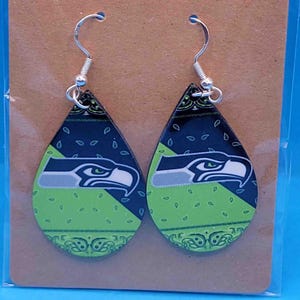 May include: A pair of teardrop earrings featuring the Seattle Seahawks logo. The earrings have a green and navy blue paisley pattern with a silver fish hook. The earrings are displayed on a brown card.