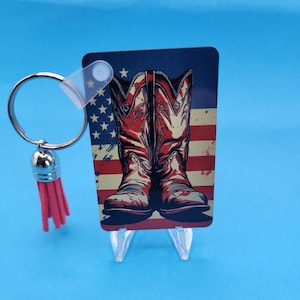 May include: A red, white, and blue American flag with a pair of red cowboy boots on top. The boots are distressed and have a vintage look. The image is printed on a clear acrylic keychain with a silver ring and a pink tassel.