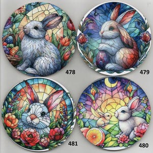May include: Four round ceramic plates with a stained glass design featuring white rabbits in different poses. Each plate has a different background color and design, including flowers, a basket of eggs, a crescent moon, and a sunrise.