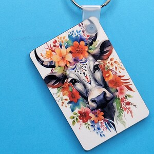 May include: A white cow with black markings and a floral crown of orange, red, pink, and blue flowers. The cow's face is the focus of the image, with its large, dark eyes and black nose.