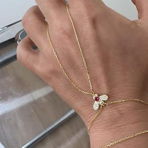 May include: A gold necklace and bracelet set. The necklace has a bee pendant with red and white gemstones. The bracelet is a simple chain. The jewellery is worn on a wrist. Jewellery.