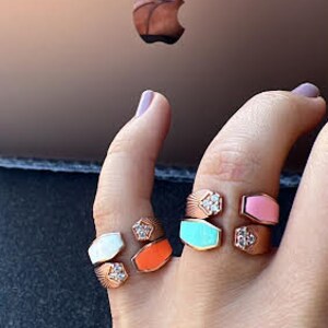 May include: A hand model wearing four different colored rings. The rings are all gold and have a geometric design. The colors of the rings are white, orange, turquoise, and pink.