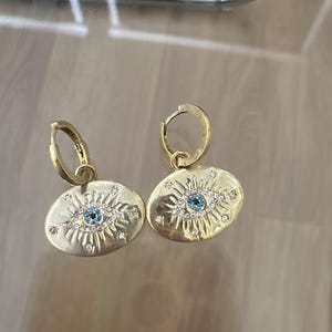 May include: A pair of gold earrings with a blue evil eye charm. The charms are oval shaped and have a blue stone in the center surrounded by smaller white stones.
