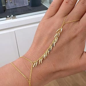 May include: Gold hand chain bracelet with a leaf design. The bracelet features a delicate chain that connects to a ring and a series of small, leaf-shaped charms. The jewelry is displayed on a hand.