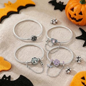 Puede incluir: Cuatro pulseras de cadena de serpiente en tono plateado con varios dijes. Uno tiene una rosa negra, otro un corazón con una piedra rosa, otro un árbol circular con piedras azules y otro una piedra morada circular con un dije de libélula.