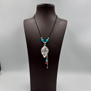 Puede incluir: Un collar con un colgante de plata de diseño geométrico y cuentas turquesa y rojas. El collar está en un cordón negro y se muestra en un maniquí negro.