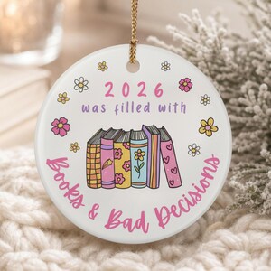 Può includere: Un ornamento rotondo in ceramica bianca appeso a un cordino dorato. Il testo 2026 was filled with Books & Bad Decisions è stampato in rosa e viola. Al centro si trova una fila di cinque libri illustrati colorati con motivi floreali.