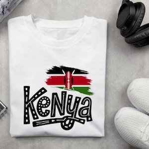 May include: White t-shirt with a graphic design featuring the word "Kenya" in a decorative font, alongside a stylized representation of the Kenyan flag. The shirt is folded on a grey surface.