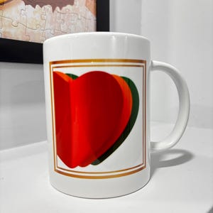 Puede incluir: Taza de cerámica blanca con un diseño de corazones rojos, naranjas y verdes superpuestos dentro de un rectángulo con borde dorado. La taza tiene un asa curva. Fondo blanco.