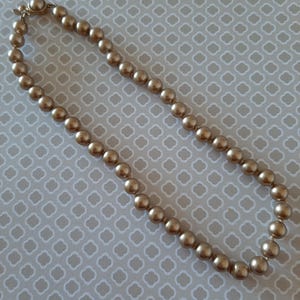 May include: A gold-colored beaded necklace with a clasp. The necklace is composed of evenly spaced, round beads. The beads have a metallic sheen and are strung together to form a complete circle. The necklace is displayed on a patterned surface.