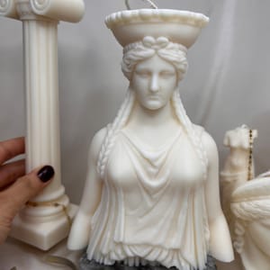 May include: White candle sculpture of a woman's bust with a crown, on a marble base. The sculpture is a replica of a classical Greek statue.