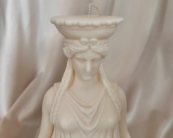 Puede incluir: Una vela blanca con forma de busto de mujer con un peinado trenzado y una prenda drapeada. La vela tiene una mecha y una parte superior decorativa.