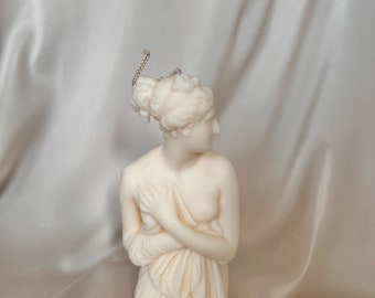 Puede incluir: Un busto de mujer de cerámica blanca con un paño drapeado, esculpido en estilo clásico. El busto es una vela y tiene una mecha.