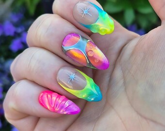 May include: A hand displaying a set of colorful, artistic nail designs. The nails feature a variety of colors, including pink, yellow, blue, and iridescent hues. Some nails have a star design, while others have a textured, bubble-like appearance.