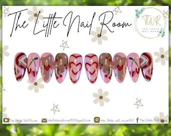 May include: A set of ten pink and white false nails with cherry and heart designs. The nails are decorated with red, white, and brown paint. The nails are arranged in a row on a white background.