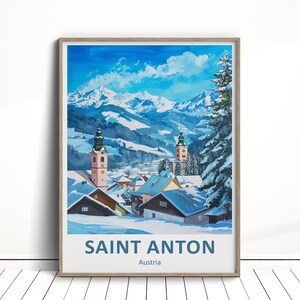 May include: A framed print of a winter landscape in Saint Anton, Austria. The artwork depicts a snow-covered village nestled in a valley, with church steeples and snow-capped mountains under a bright blue sky. The text "SAINT ANTON" is at the bottom.