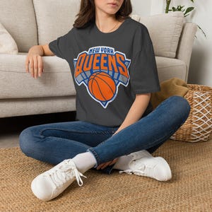 May include: A dark gray short-sleeve t-shirt featuring a graphic design with the text NEW YORK QUEENS in orange and white lettering above an orange basketball illustration with a blue and white outline, paired with blue denim jeans and white sneakers.