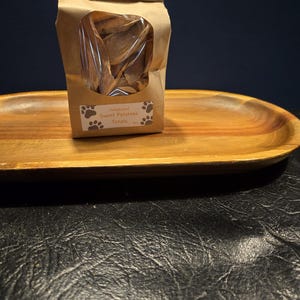 May include: A brown paper bag with a clear window, filled with dried sweet potato treats for dogs. The bag has a label with paw prints and the words "Sweet Potatoes Treats". The bag sits on a wooden oval tray.