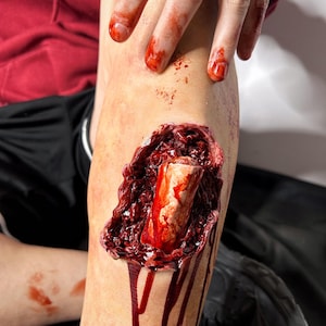 May include: A gruesome special effects prosthetic of an open wound on a leg, revealing bone and tissue. The wound is surrounded by dark red blood and gore, with blood dripping down the leg. A hand with blood-stained fingers is visible.