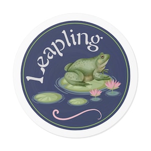 Puede incluir: Una pegatina circular azul y blanca con la palabra "Leapling" escrita en cursiva blanca. Una rana verde se sienta en un nenúfar con flores rosas en el agua.