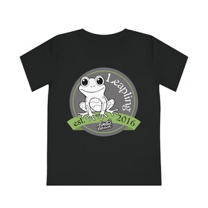 Puede incluir: Camiseta negra con un gráfico circular verde y gris que presenta una rana de dibujos animados y el texto "Leapling est. 2016 Edición limitada".