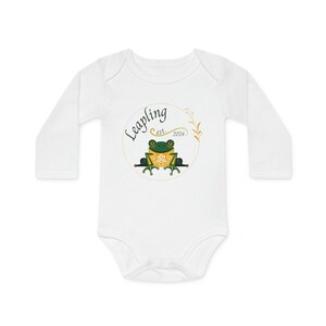 Puede incluir: Body blanco de manga larga para bebé con una rana verde sentada en una rama dentro de un círculo dorado con el texto "Leapling est. 2024"