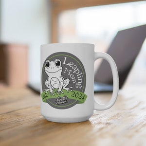 Puede incluir: Taza de cerámica blanca con un diseño gris y verde que presenta una rana de dibujos animados y el texto "Leaping Mom est. 2024 edición limitada".