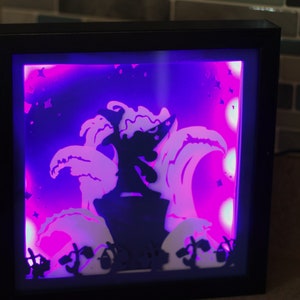 May include: A black framed shadow box with a purple and white light inside. The shadow box features a silhouette of a figure standing on a wave with stars in the background.