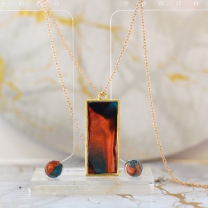 May include: A gold-toned necklace with a rectangular pendant featuring a blend of red, orange, and teal colors. The pendant is framed in gold and hangs from a delicate chain. Two matching round earrings are also visible.