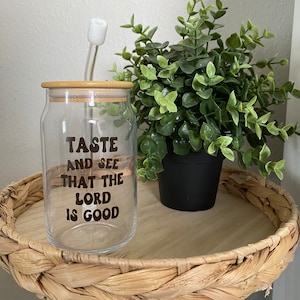 May include: A clear glass tumbler with a bamboo lid and a glass straw. The tumbler has the text "TASTE AND SEE THAT THE LORD IS GOOD" in brown lettering. A small green plant in a black pot sits next to the tumbler.