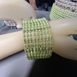 May include: A light green beaded bracelet with multiple strands of small, round beads. The bracelet is displayed on a mannequin arm. Another bracelet with white and green beads is visible in the background.
