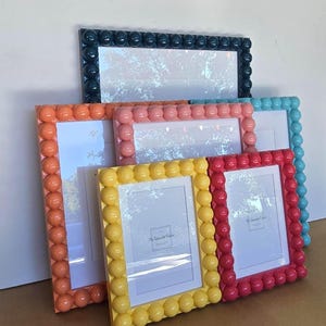 May include: A collection of colorful picture frames. The frames are rectangular with a border of round, bubble-like embellishments. Colors include navy, orange, pink, turquoise, yellow, and red. The frames are empty, with white backing and glass.