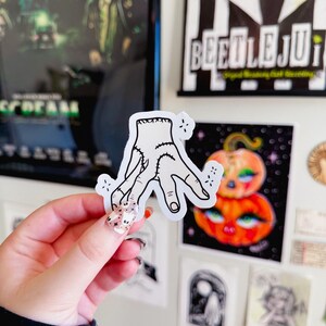 May include: A white sticker featuring a hand with stitches, a design inspired by the Addams Family. The sticker is held in front of a wall decorated with posters, including a Beetlejuice poster and a pumpkin illustration.