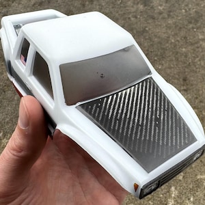 May include: A white toy truck body with a carbon fiber hood. The truck has a clear windshield and side windows. An American flag sticker is visible on the side window. The truck is held in a person's hand.