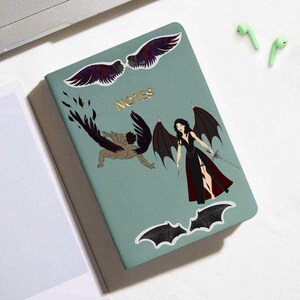 Puede incluir: Una libreta verde con la palabra "NOTES" escrita en letras doradas. La libreta tiene varias pegatinas, incluyendo un ángel negro y rojo con alas, un ángel negro y blanco con alas y un murciélago negro.