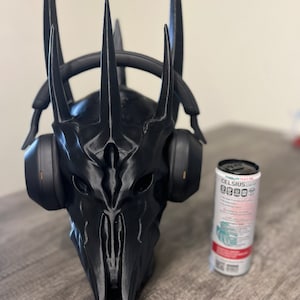 May include: A black 3D printed helmet with a crown of spikes, resembling a character from a fantasy movie, is worn by a pair of black headphones. The helmet is sitting on a wooden surface next to a can of Celsius energy drink.