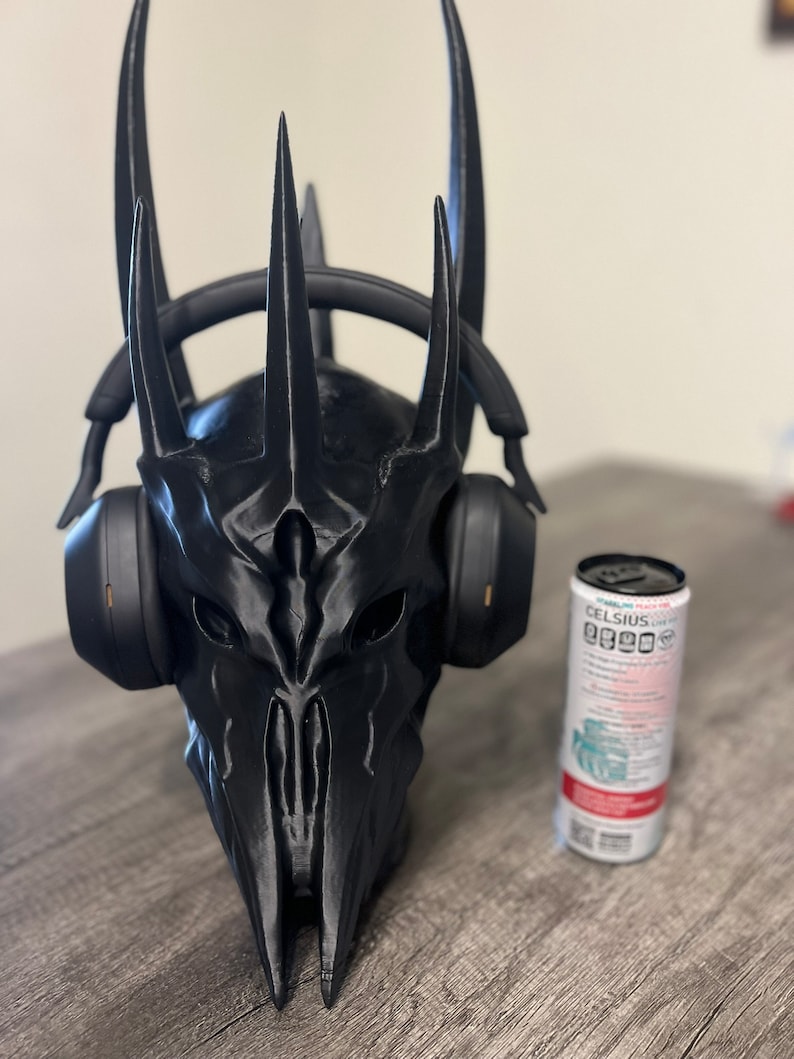 May include: A black 3D printed helmet with a crown of spikes, resembling a character from a fantasy movie, is worn by a pair of black headphones. The helmet is sitting on a wooden surface next to a can of Celsius energy drink.