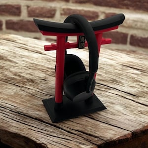May include: Black headphones resting on a red and black torii gate-shaped stand. The stand has a black base and the torii gate is made of red plastic. The stand is sitting on a wooden table.