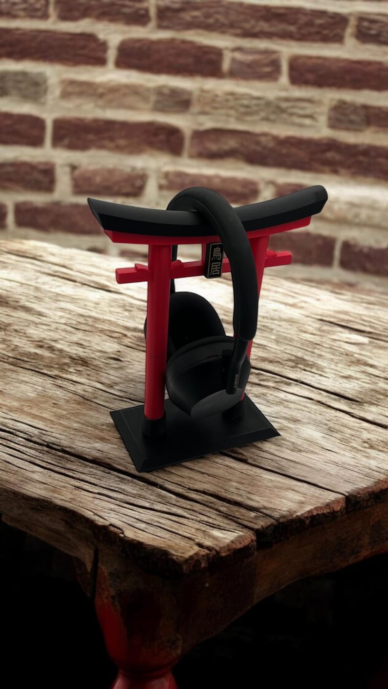May include: Black headphones resting on a red and black torii gate-shaped stand. The stand has a black base and the torii gate is made of red plastic. The stand is sitting on a wooden table.