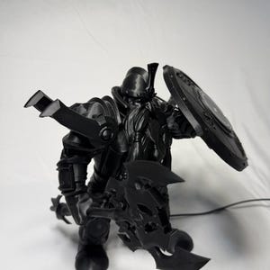 May include: A black 3D printed figurine of a warrior with a long braided beard, wearing heavy plate armor and a helmet. The figure holds a large ornate battle axe in one hand and a circular shield in the other, with prominent shoulder pauldrons.