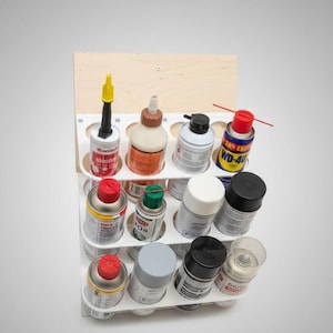 May include: A white, multi-tiered organizer holding various spray cans and bottles. The organizer is mounted on a light-colored wooden backboard. The cans and bottles are of different sizes, colors, and brands, including WD-40.
