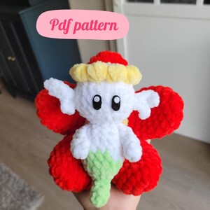 May include: A crocheted plush toy in the shape of a red flower with a white character in the center. The character has a yellow crown and a green tail.