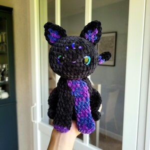 Könnte beinhalten: Ein schwarzes, gehäkeltes Plüschkatzenspielzeug mit violetten und blauen Garn-Akzenten. Die Katze hat glitzernde grüne Augen und ein Halbmond-Design auf der Stirn.