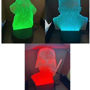 Pode incluir: Três lâmpadas LED com silhuetas de personagens em acrílico. Uma lâmpada brilha em verde com uma figura de Yoda, uma em ciano com uma figura de Darth Maul e uma em vermelho com uma figura de Darth Vader. Cada lâmpada tem uma base preta circular.