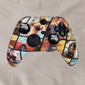 May include: A wireless video game controller featuring a multi-panel graphic print of Grand Theft Auto characters and scenes. The controller includes black analogue sticks with Grand Theft Auto logos, a black directional pad, and four coloured action buttons.