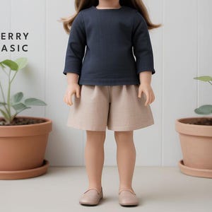 Op de afbeelding: Een pop die een marineblauw shirt met lange mouwen en een beige broek draagt. De pop staat voor een witte muur met twee planten in potten.